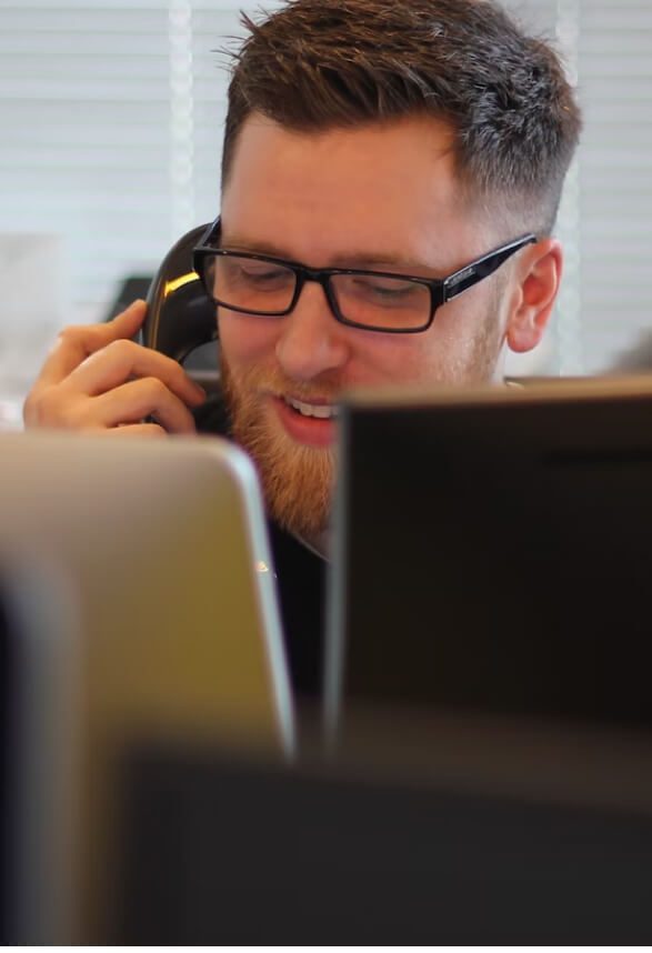 A man is sitting at his desk, on the telephone. We can see his face, but it is partially covered by two computer monitors. A man is sitting at his desk, on the telephone. We can see his face, but it is partially covered by two computer monitors.