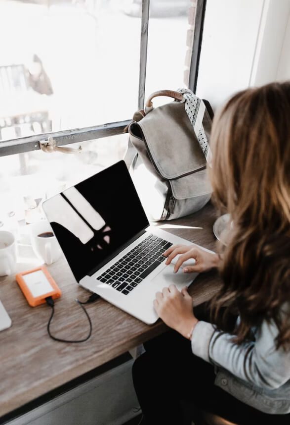 A woman sits at a desk nook facing a window. She is looking at something not visible on her laptop screen.