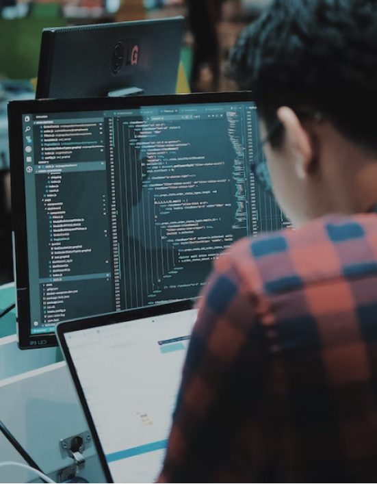 A man works on his laptop computer screen. He has a larger desktop monitor, and both screens explore code.