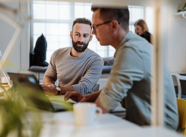 Developer with beard leaning over talking to another developer in the office.