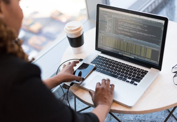 A woman sits at her laptop looking at code on her screen. Her left hand is reaching for her mobile telephone.